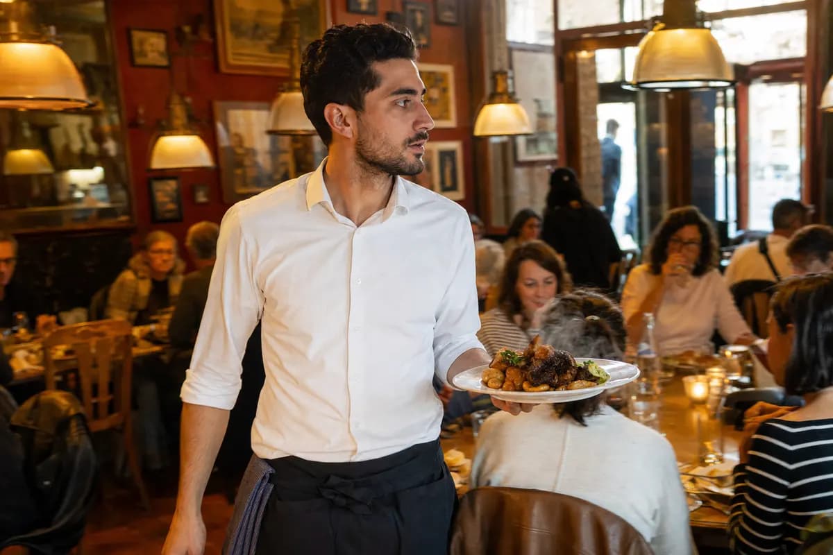 Servicemitarbeiter trägt einen frisch angerichteten Teller durch ein gut besuchtes Restaurant im warmen Abendlicht